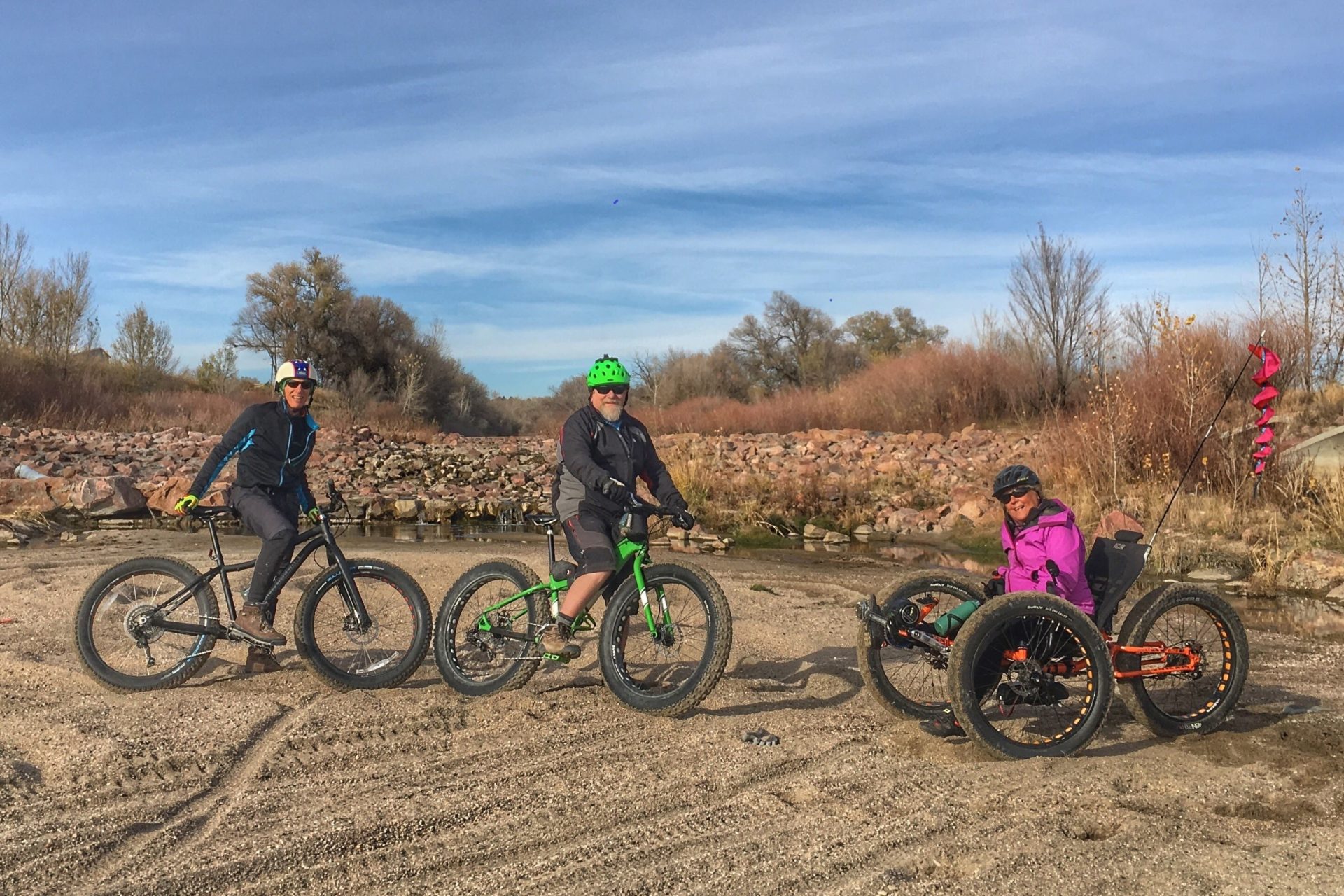 Three people pose for the camera with their bikes standing on a dirt patch. The person on the right is sitting in an off-road trike.