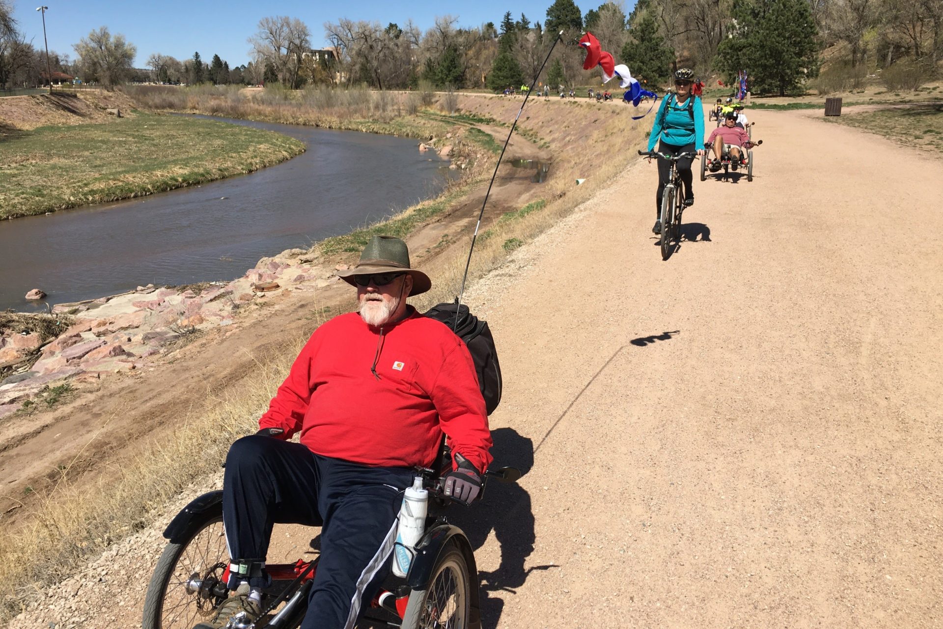 People ride on a dirt trail. In the foreground is a person riding a recumbent trike. Further in the background there are more people riding recumbent trikes.