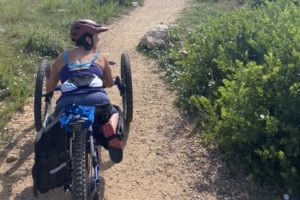 Quinn Brett rides a prone trike on a dirt trail. She is facing toward the horizon.