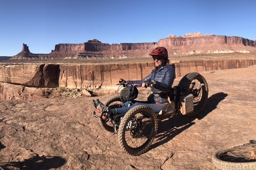 A person poses in a tadpole upright trike on red rock in front of a red canyon.
