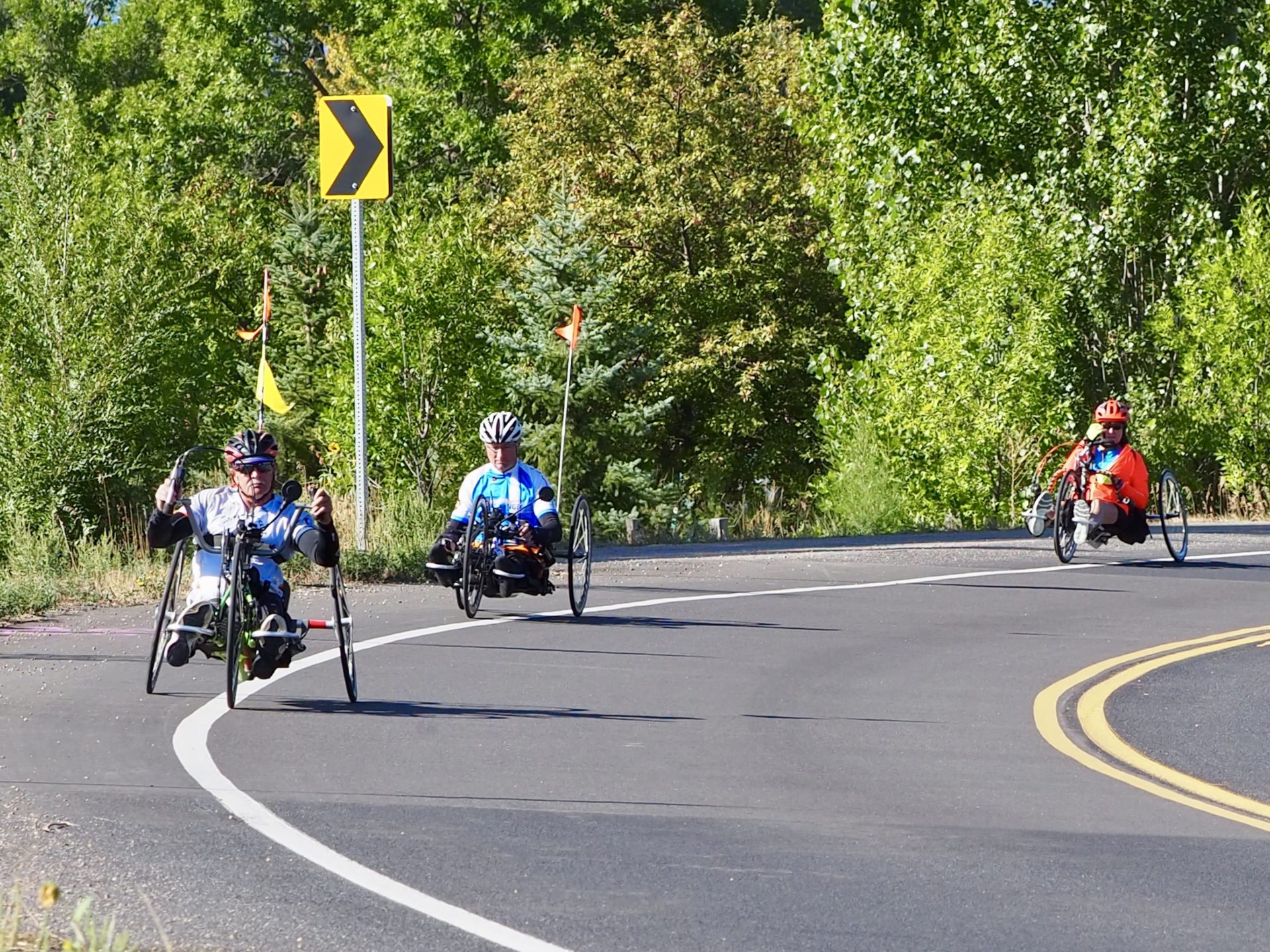 Three people ride recumbent trikes on a paved road. There are green trees behind them.