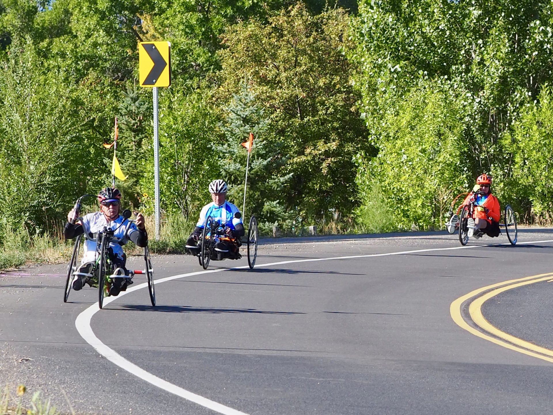 Pedal 4 Possible 9-14-19 Three people ride recumbent trikes on a paved road. There are green trees behind them.