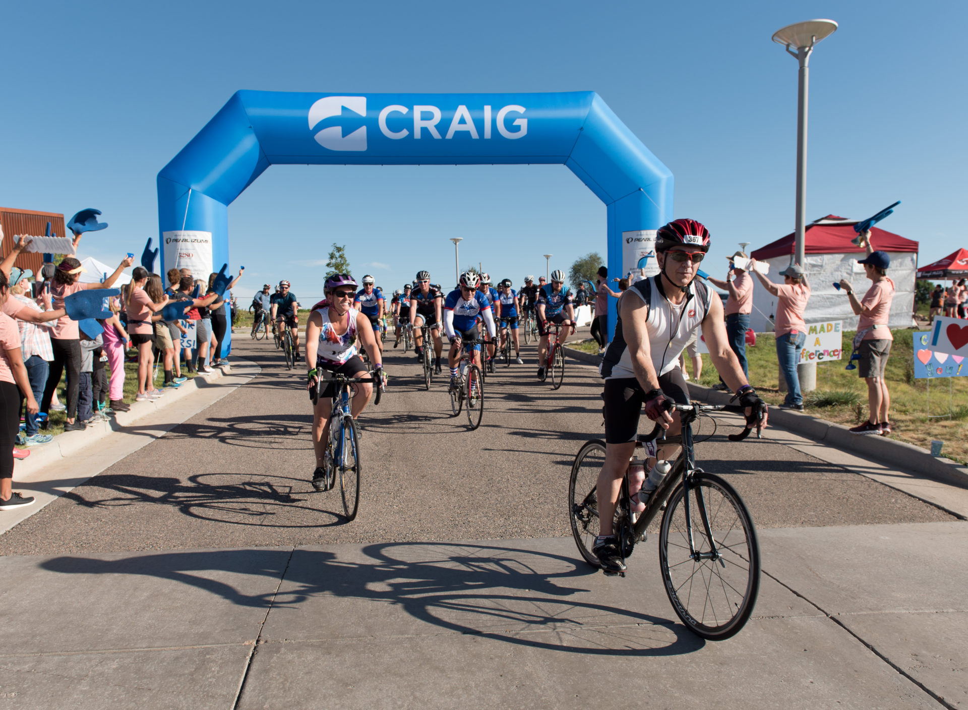 Bicyclists cross the finish line of a ride coming toward the camera. They are on a paved road. There is an inflatable arch above the road that says "Craig".