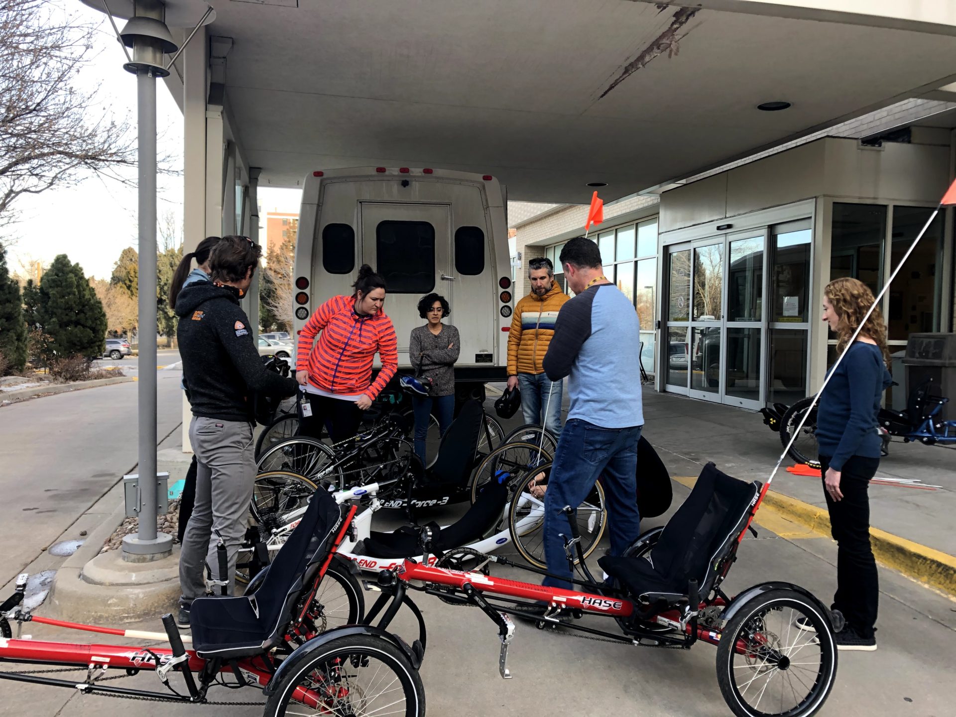 A group of people stand around a number of adaptive bikes under an awning in front of a building.