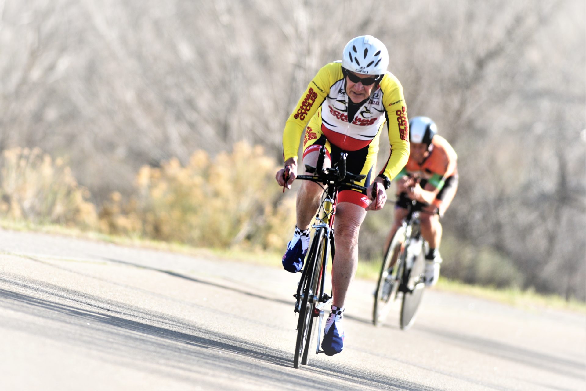 Two people ride road bikes on a paved road toward the camera. One person is in front of the other.