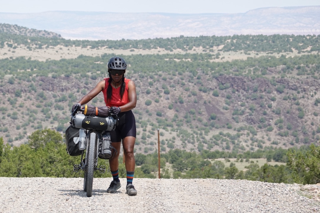 A person stands next to their bike. They are on a gravel road. Behind them in the distance are desert hills.