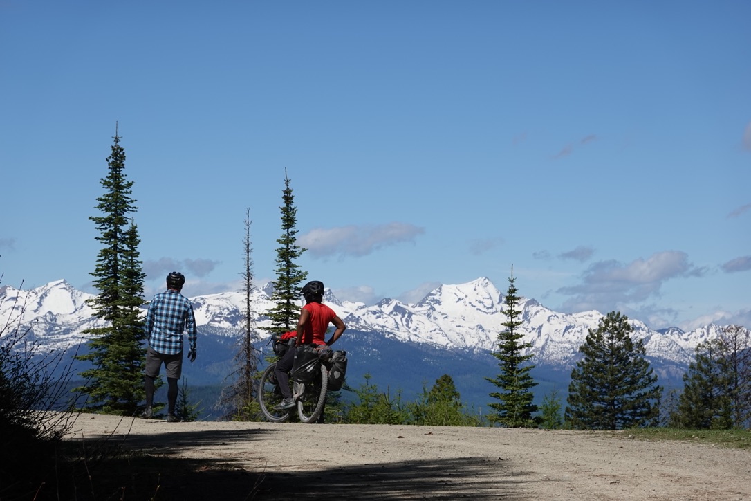 Two people stand on a dirt road facing away from the camera. One of them has a bike. They are looking toward the large mountains in the background.