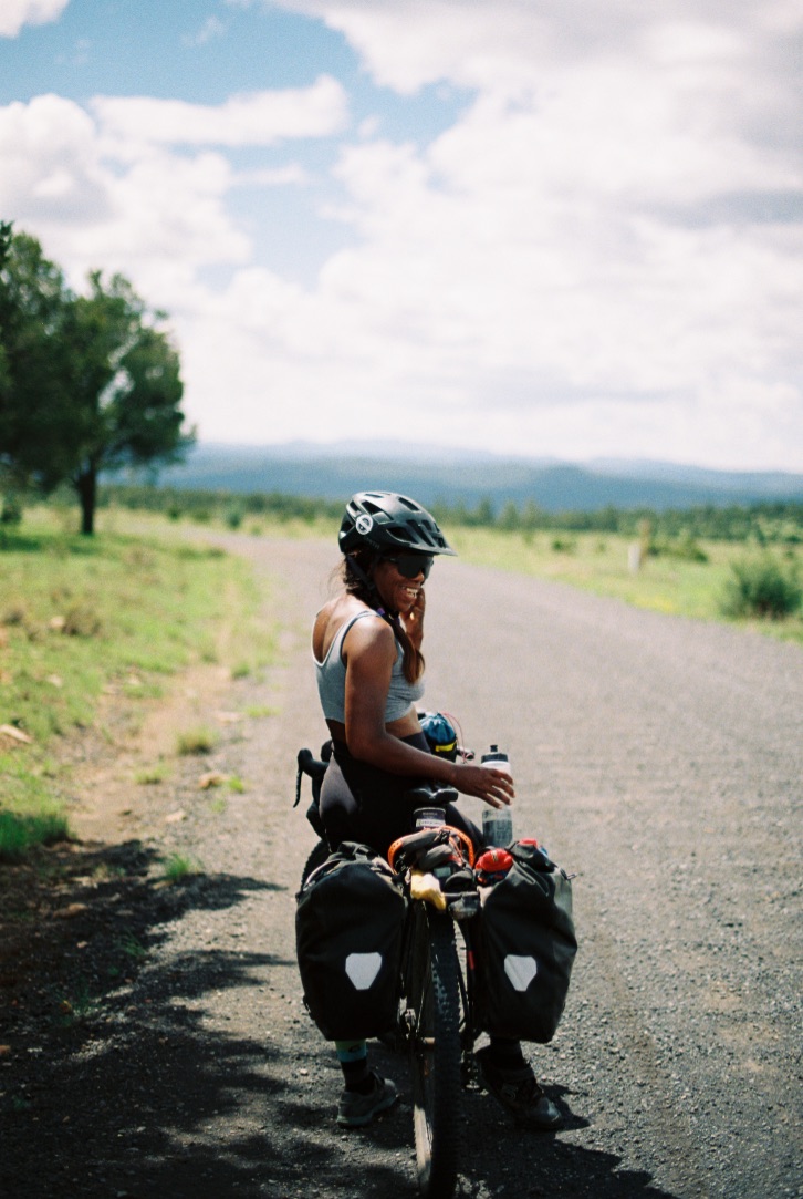 A person standing over their bike on a dirt road with their body facing away but face turned back to the camera and smiling. 