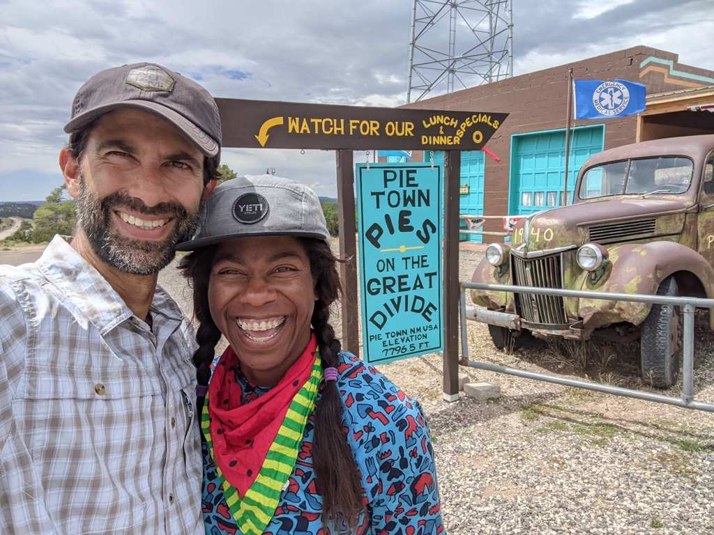 Two people take a selfie smiling together. They stand in front of a sign that reads "Pie Town Pies on the Great Divide."