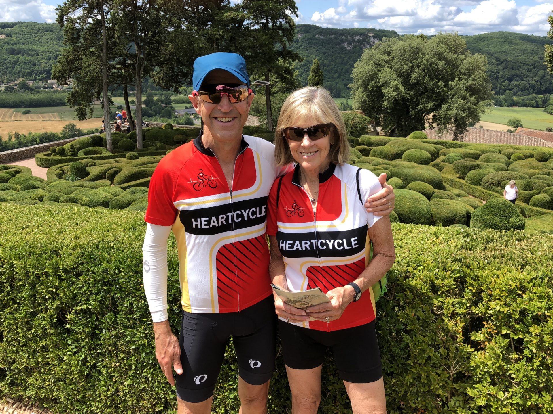 A white man and woman pose wearing cycling jerseys and sunglasses. They are photographed from the knees up. They are standing in front of shrubs.