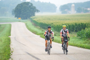 2 bicyclists smile as they bike on a back country road through lush, foggy fields and trees