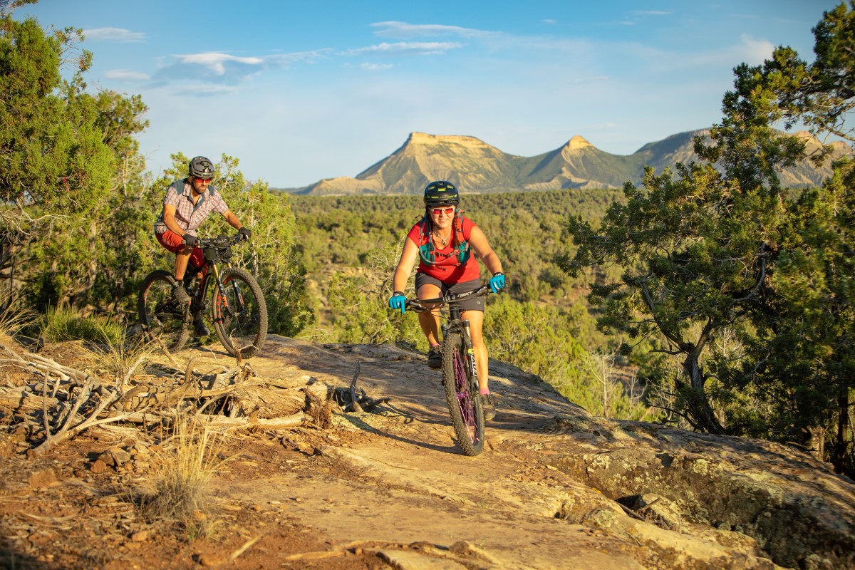 Two people riding mountain bikes on a dirt trail toward the camera, with a valley and mountains behind them off the edge of the trail.