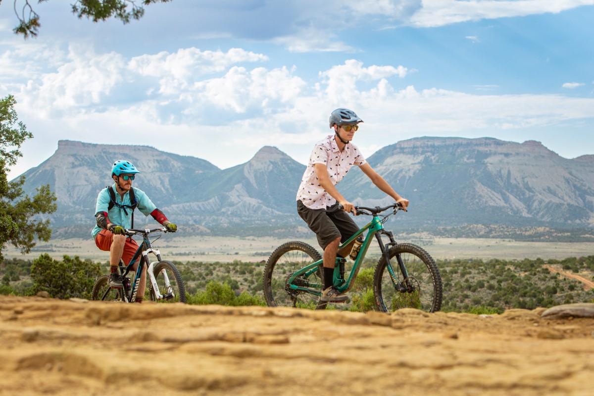 Two people ride mountain bikes toward the left of the frame, with a valley and mountains in the background.