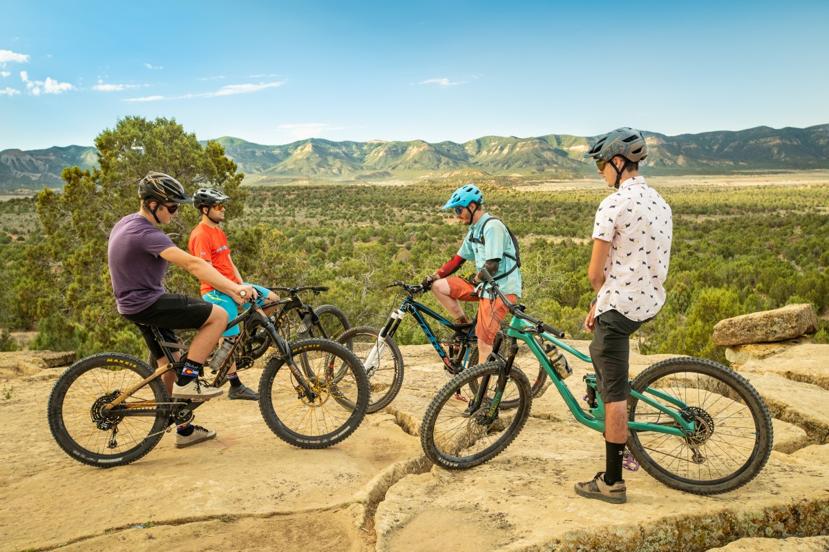A group of four people stand with mountain bikes on rocks, above a valley of brush with hills in the far background.