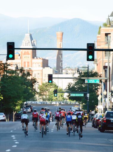 People riding bikes away from the camera on a street, passing stoplights with the Coors factory in the background and mountains in the far background.