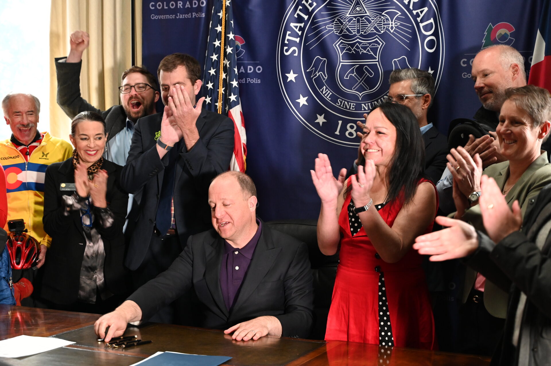 The Governor of Colorado signing a bill at his desk surrounded by people standing and cheering.