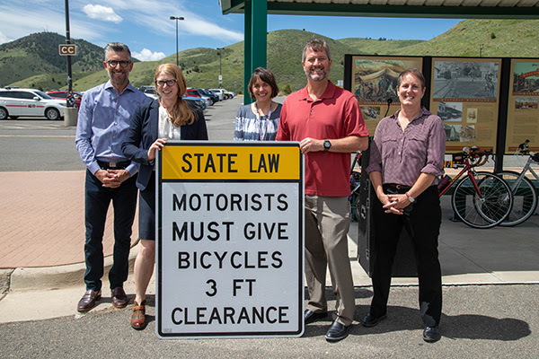 Five people stand and smile in next to a sign that says "State law: Motorists must give bicyclists 3 ft clearance."