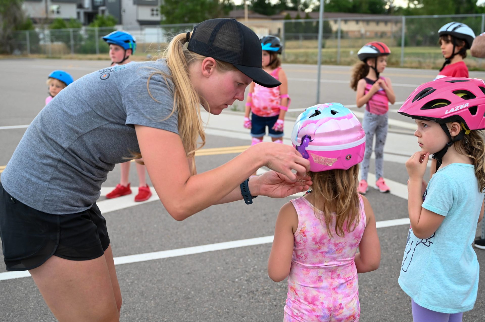 A woman leans down to adjust the bike helmet of a small child facing away from the camera, with several other children standing nearby.