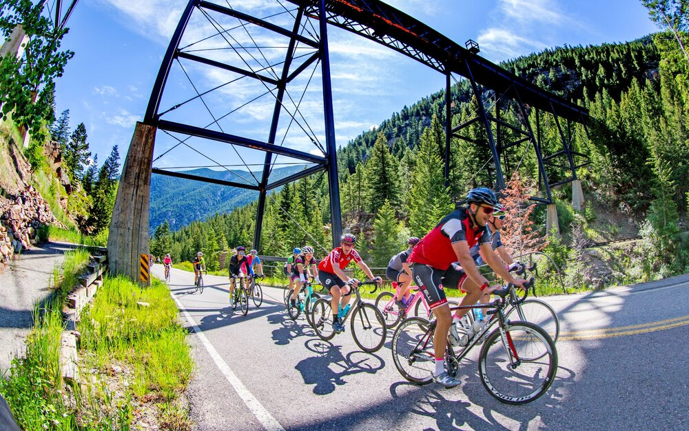 a large group of cyclist ride on the road, with a train bridge and the mountains in the background, during the Triple Bypass.