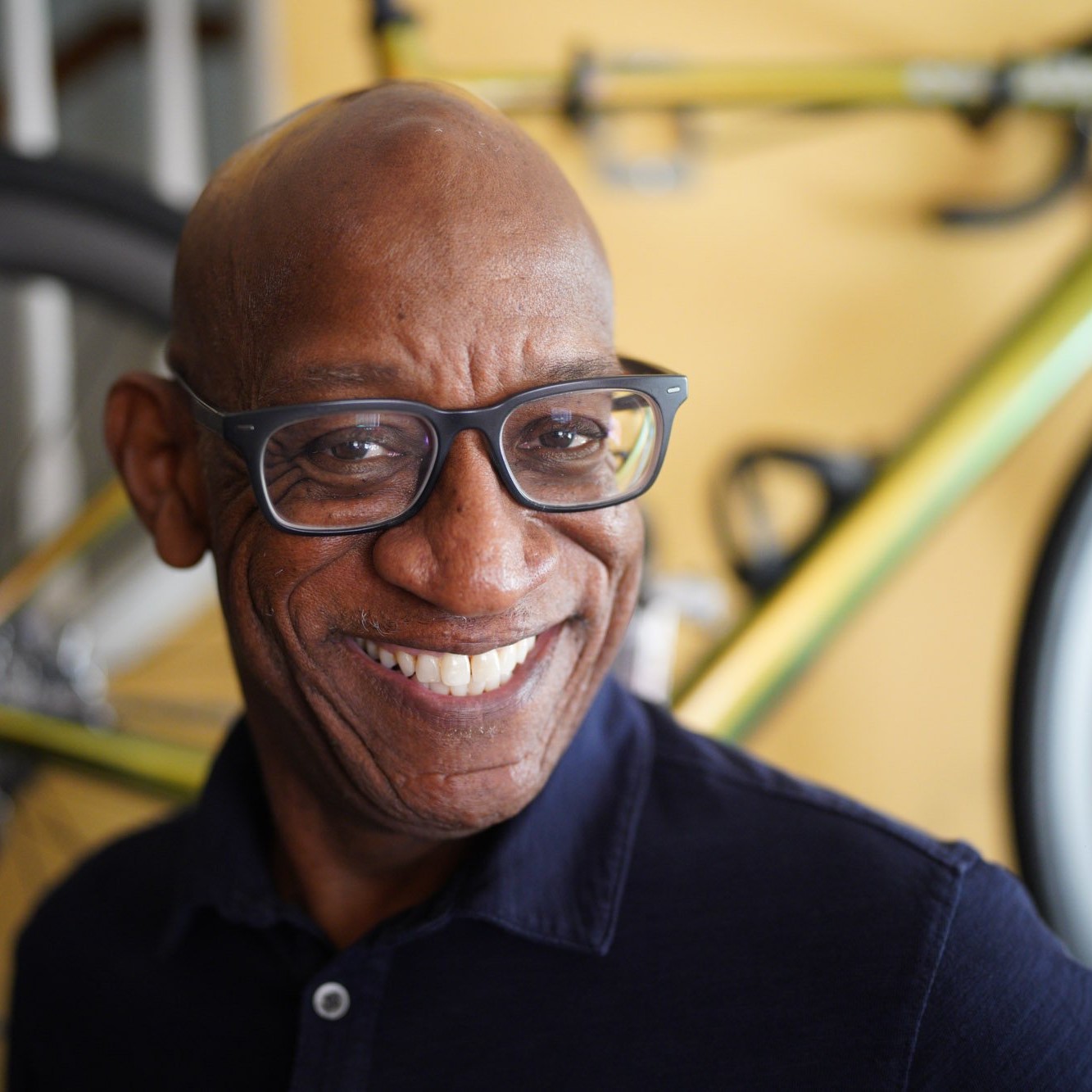A Black man wearing glasses, photographed from the chest up with a bike in the background.