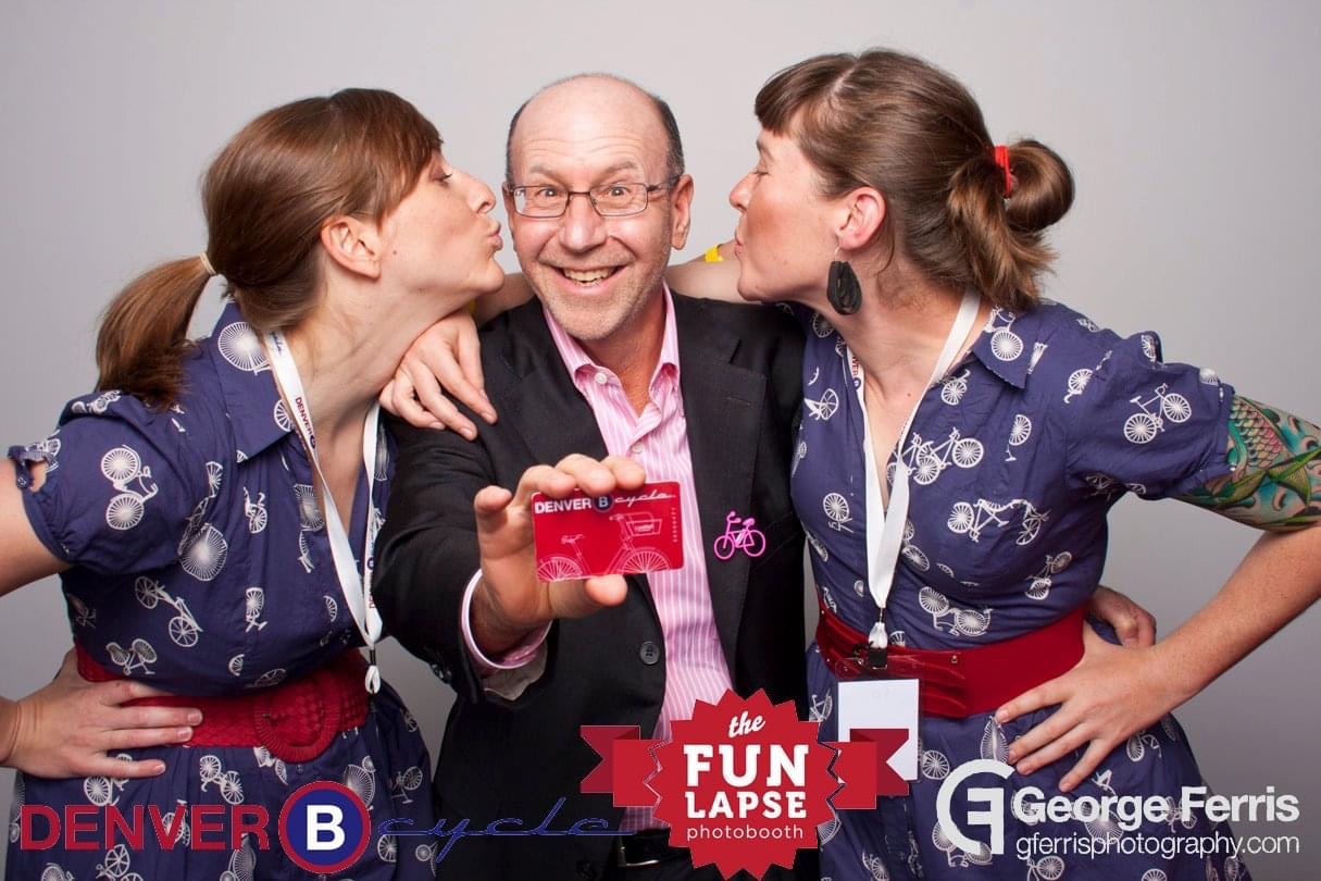 Emily Snyder and Maggie Thompson, wearing bicycle-printed dresses, kiss Steve Sander on the cheek as he holds a Denver B-Cycle membership card